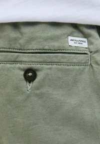 Tela verde claro con un acabado texturizado, que presenta un botón negro y una etiqueta que dice "JACK & JONES EST.1990" en la cintura.
