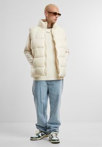 Puffy cream vest with high collar, worn over a light shirt. Light blue jeans and green-and-white sneakers. Minimalist design, smooth texture.
