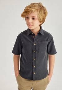 Camisa de manga corta gris oscuro con cuello, presenta un bolsillo en el pecho y botones marrones, hecha de una tela suave.