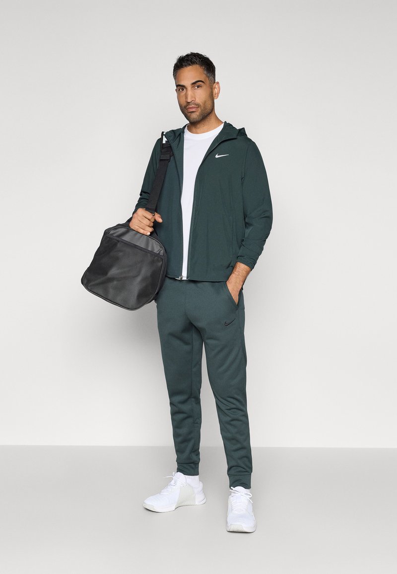 Chaqueta deportiva verde oscura con capucha y logo, camiseta blanca, pantalones jogger a juego y zapatillas blancas. Lleva una bolsa negra con una superficie texturizada.
