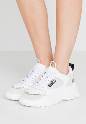 Hvide chunky sneakers med grå accenter og Versace Jeans Couture-mærke båret på bare fødder mod en ensfarvet baggrund.