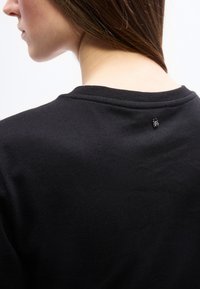 Νεαρή γυναίκα με καστανά μαλλιά που φοράει μαύρο τοπ, εμφανιζόμενη από πίσω, εστιάζοντας στη λαιμόκοψη και στη μικρή μεταλλική λεπτομέρεια κοντά στο γιακά.