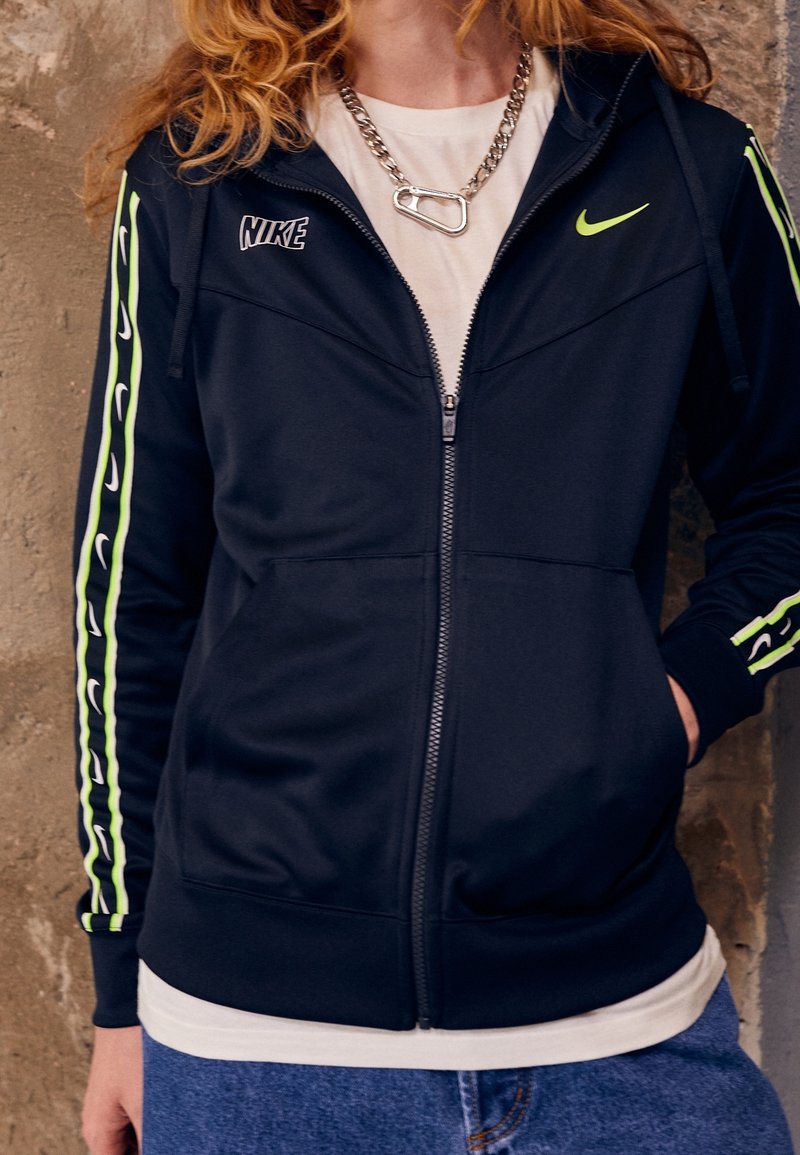 Modrá mikina Nike na zip s neonově zelenými pruhy po stranách. Má plný přední zip, kapsy a lehkou, hladkou látku. Viditelný řetízkový doplněk.