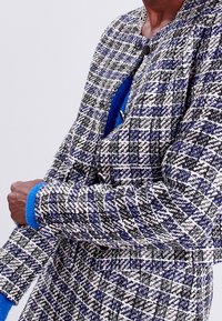 Tweed jacket with a checkered pattern in black, white, and blue, featuring a round black button and plush blue cuffs. Structured shape.