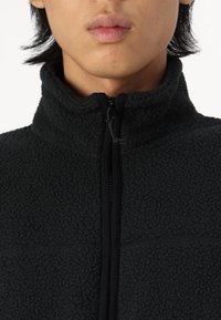 Sort fleecejakke med høj krave og frontlynlås. Har en blød tekstur og kontrastende sorte lynlåse. Minimalistisk design.