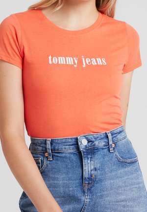 Femme portant un t-shirt orange ajusté avec le texte « tommy jeans », rentré dans un jean en denim bleu taille haute.