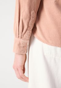 Light pink corduroy shirt with a buttoned cuff. Features vertical ribbed texture and a rounded hem, contrasting with a white garment underneath.