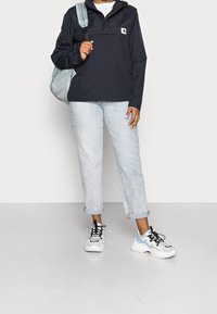 Navy windbreaker with a hood, front pocket, and logo patch. Light blue jeans with a slight wash. Chunky white and blue sneakers with black accents.