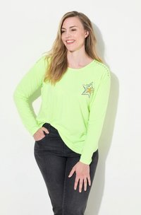 Langärmliges, hellgrünes Baumwollhemd mit einem Stern-Grafikdesign und orangefarbenen Akzenten sowie dekorativer Stickerei an den Schultern. Kombiniert mit dunklen Jeans.