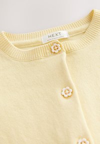 Lysegul cardigan med ribbet halsudskæring, der har to hvide knapper formet som blomster og en blød tekstiltekstur. Labelen siger "NEXT."