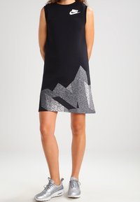 Svart ärmlös klänning med en silverfärgad bergsgraphic vid fållen. Har en Nike-logotyp i vitt på bröstet. Bärs med silverfärgade sneakers.