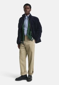 Marine blauwe gewatteerde jas over een groene cardigan, lichtblauw gestreept shirt, beige broek en zwarte veterschoenen. Gelaagde en gestructureerde outfits.
