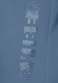 Helles blaues Hemd mit einer strukturierten Oberfläche, das auf einer Seite glitzernde Strassverzierungen in einem geometrischen Muster aufweist.