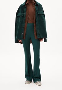 Camisa de pana verde de corte oversized, cuello alto marrón, pantalones acampanados en color verde azulado y zapatillas a rayas. Presenta bolsillos y materiales texturizados.