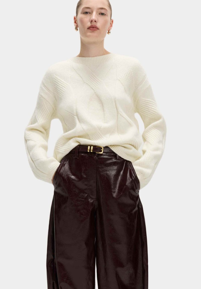Jersey de punto crema con patrones de trenzado, combinado con pantalones de imitación cuero burdeos oscuros de cintura alta, que cuentan con un cinturón con una hebilla dorada.