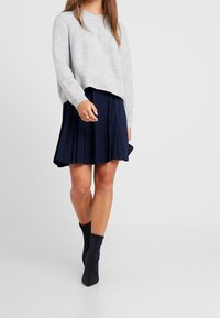 Svetle šedý pletený svetr s kulatým výstřihem, doplněný skládací tmavě modrou sukní. K outfitu se hodí černé boty po kotníky.