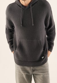 Dunkelgrauer, gerippter Hoodie mit V-Ausschnitt und Kordeln, ausgestattet mit einer Fronttasche und einem kleinen Logo auf der unteren Seite. Weicher Stoff.