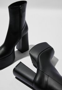 Botas de cuero negro de plataforma hasta el tobillo con tacones de bloque y cremalleras laterales, mostradas sobre una superficie blanca con detalles de sombra.