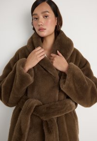 Roupão de pele sintética castanho com um colarinho largo, cintura cintada e mangas oversized, apresentando uma textura suave e aveludada em toda a peça.