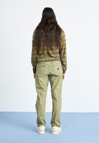 Striped long-sleeve shirt in green and orange, paired with light green cargo pants featuring back pockets and a logo patch, worn with white shoes.