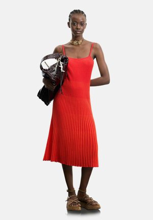 Kobieta ubrana w czerwoną prążkowaną sukienkę midi i brązowe sandały na platformie, trzymająca czarną skórzaną torbę z paskiem ozdobionym ćwiekami na ramieniu.