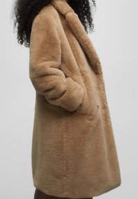 Karamelový kabát z umělé kožešiny s vysokým límcem, oversize střihem a postranními kapsami, s hladkou strukturou a minimalistickým designem.
