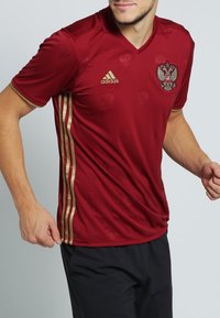 Röd idrottströja med gyllene detaljer; har en V-hals, korta ärmar och den ryska emblemet på bröstet. Texturerad tyg med subtila mönster.