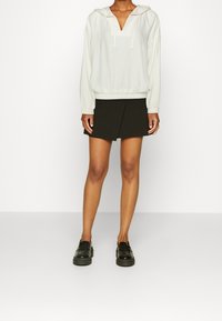 Lyscremet pullover med hætte og V-hals, parret med en sort omslagsnederdel. Modellen har sorte loafers på og står foran en neutral baggrund.