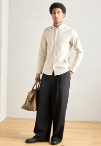 Μπεζ πουκάμισο με κουμπιά, μαύρο παντελόνι με φαρδιά πόδια και μαύρα παπούτσια. Καφέ τσάντα με χερούλια. Λείο ύφασμα και εφαρμοστή γραμμή. Ουδέτερο περιβάλλον.