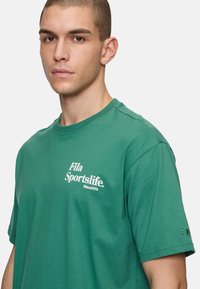 Camiseta de algodón verde con mangas cortas, cuello redondo y logo blanco "Fila Sportslife" en el pecho, además de un pequeño logo en la manga.