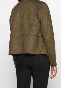 Giacca in suede marrone con design aderente, cucitura dritta sul retro e orlo leggermente svasato. Presenta dettagli cuciti e maniche lunghe.