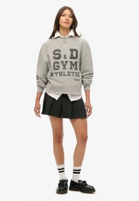 Grijze sweater met zwarte letters, gelaagd over een wit shirt, gecombineerd met een zwarte geplooide rok, witte sokken met zwarte strepen en zwarte schoenen.