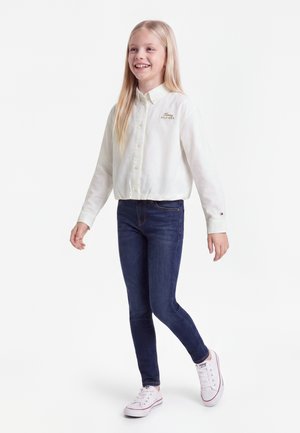 Fille souriante aux longs cheveux blonds portant une chemise blanche boutonnée, un jean bleu foncé et des baskets blanches, marchant sur un fond blanc.