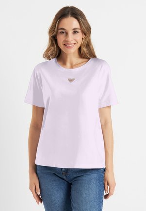 Glimlachende vrouw met schouderlang haar, gekleed in een lichtpaarse shirt met korte mouwen en een klein hartvormig uitgesneden detail op de borst, en een blauwe spijkerbroek.