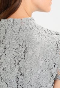 Vaaleanharmaa pitsitoppi, jossa on kukkakuosi, läpikuultava rakenne ja korkea kaula-aukko. Takana on pieni kiinnitysdetaali.