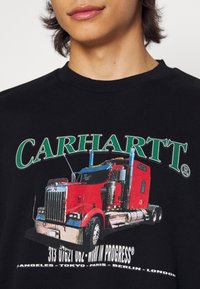 Camiseta negra con un gráfico de un camión semirremolque rojo con detalles en plata, debajo del logo de "CARHARTT" en verde y texto descriptivo debajo.