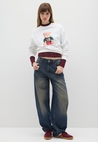 Jonge vrouw met lang haar, gekleed in een witte teddybeer-trui, een bordeauxrode kanten top, wijde vervaagde jeans en bordeauxrode sneakers, staand met haar handen in de zakken.