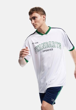 Młody mężczyzna w białej i zielonej sportowej koszulce oraz granatowych krótkich spodenkach wygląda, jakby biegł lub pozował w ruchu na jednolitym tle.