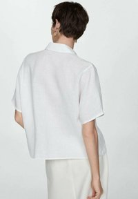 Short-sleeve white linen shirt with a collar, relaxed fit, visible textured fabric, and a slightly curved hem. Simple design with no patterns.