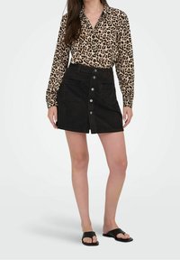 Overhemd met luipaardprint en lange mouwen, gecombineerd met een zwarte corduroy minirok met knoopsluiting aan de voorkant en zakken. Zwarte sandalen.