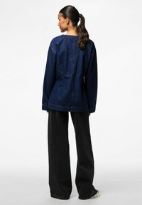 Tamno plavi traper jakna s opuštenim krojem, s vidljivim šavovima i dugim rukavima, u paru s širokim crnim hlačama.