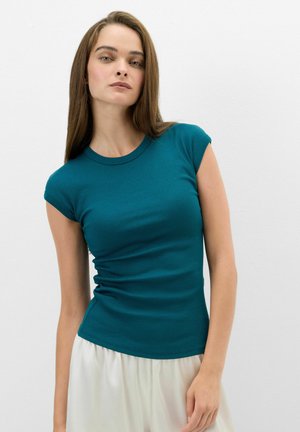 Jeune femme aux longs cheveux bruns portant un haut ajusté à manches courtes bleu sarcelle et un pantalon blanc sur un fond blanc uni.