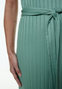 Helles grünes, geripptes Kleid mit einem gebundenen Gürtel in der Taille, vertikalen Streifen und einem weichen, strukturierten Stoff. Eine Hand ruht auf dem Kleidungsstück.