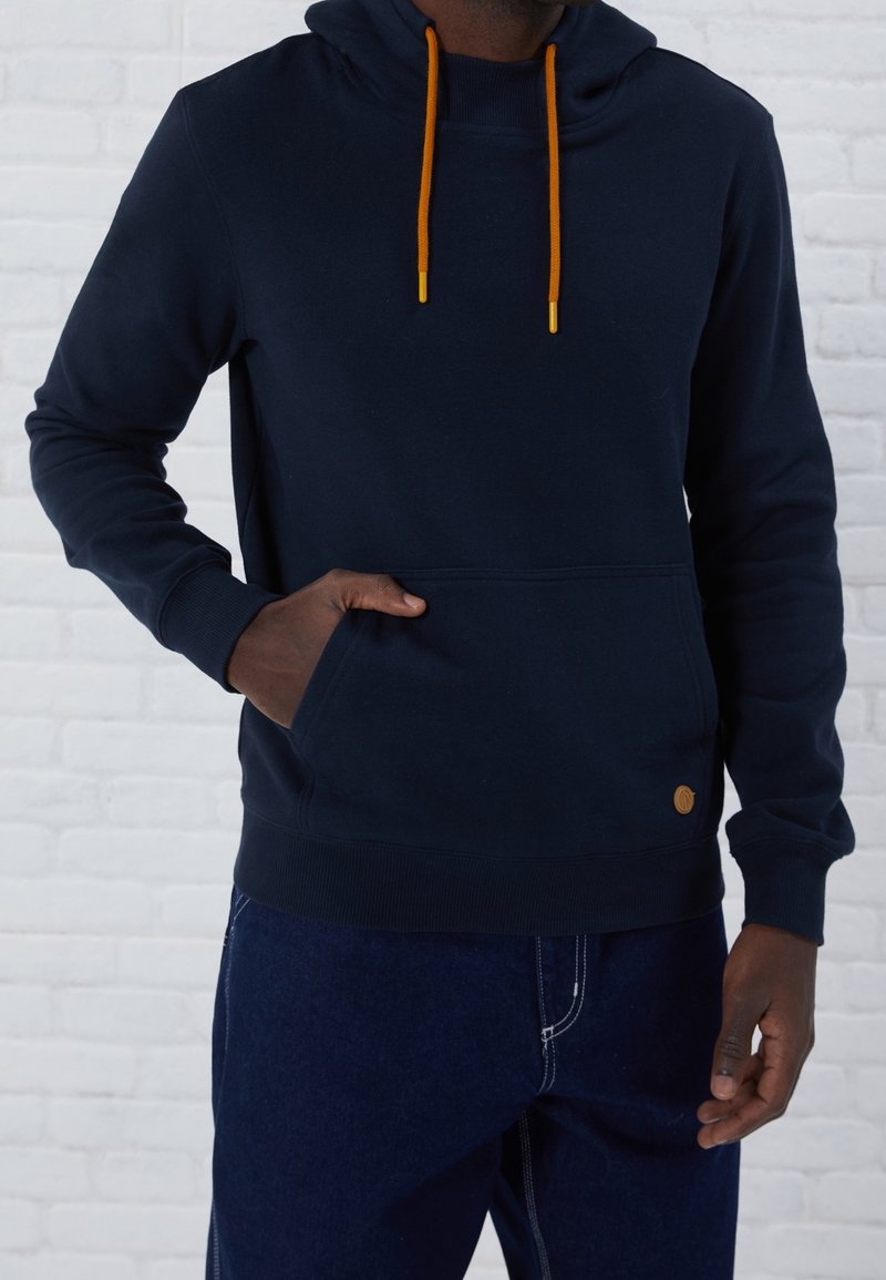 Homme portant un sweat à capuche bleu marine avec cordons orange et un jean bleu foncé, main partiellement dans la poche du hoodie devant un mur en briques blanches.