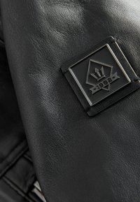 Chaqueta de piel negra con un acabado texturizado y un parche de logo metálico con "1982" en el lado izquierdo.