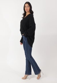 Suéter negro de punto oversized con un escote en V profundo, combinado con jeans de campana azul y sandalias de tacón transparente. Textura suave y ajuste relajado.