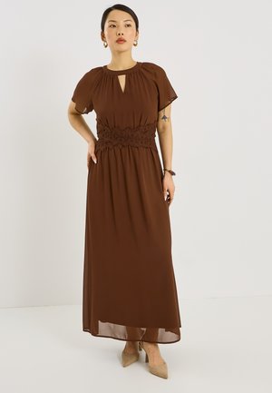 Mujer con vestidomaxi marrón con detalles de encaje en la cintura, mangas cortas y tacones beige, posando frente a un fondo claro liso.