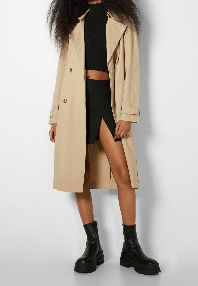 Femme portant un trench-coat beige sur un top court noir, une jupe noire fendue haute et des bottines noires à plateforme, debout contre un mur blanc.