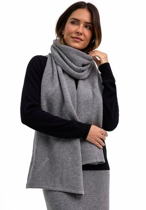 Femme portant un haut long noir avec une écharpe en tricot grise enroulée autour du cou et une jupe assortie, regardant légèrement vers la droite.