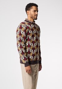 Maglione lavorato a maglia con un motivo floreale in bordeaux, crema e oro, con polsini a coste e un colletto a contrasto. Indossato con pantaloni beige.
